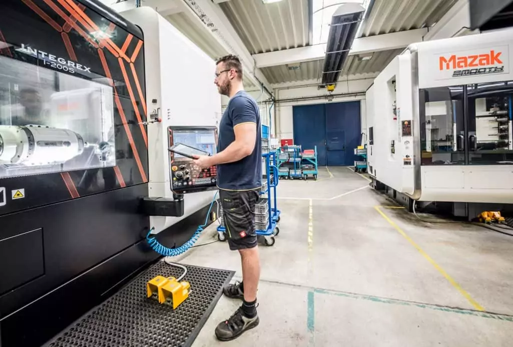 man wearing shorts watching a Mazak CNC machine holding a pendant in a factory