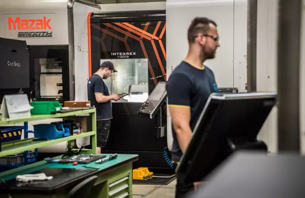 man wearing shorts watching a Mazak CNC machine holding a pendant in a factory